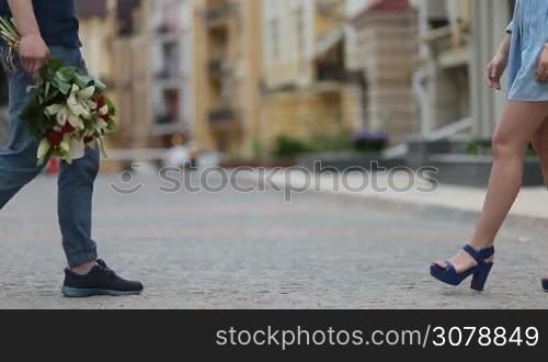 Low section of male and female legs in casual footwear meeting on romantic date over urbanscape background. Dating couple in love holding hands. Affectionate hipster man holding bunch of flowers to surprise his beloved girlfriend on a date. Side view