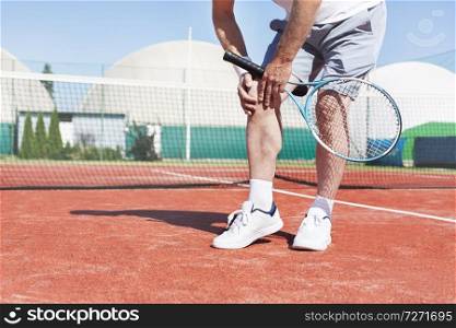Low section of mature man holding tennis racket while suffering from knee pain on red tennis court during summer
