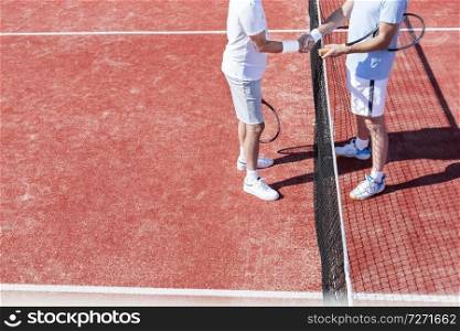 Low section of men shaking hands while standing by tennis net on red court during match