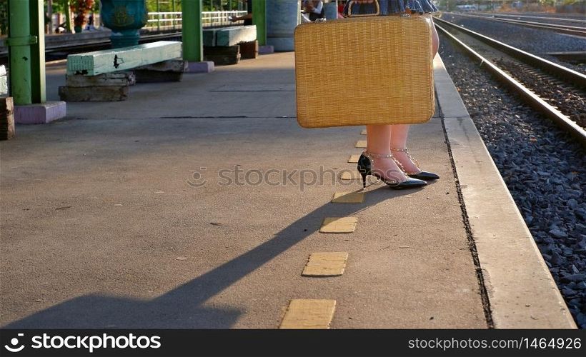 Low section of young female traveler holding vintage bamboo suitcase and waiting for train with sunlight and shadow on platform at railway station in evening time