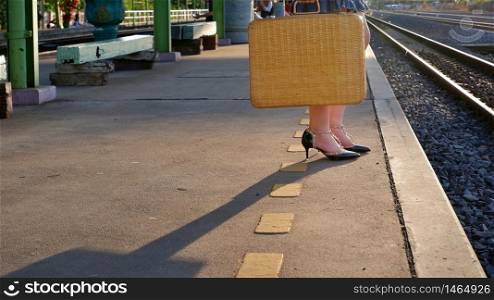 Low section of young female traveler holding vintage bamboo suitcase and waiting for train with sunlight and shadow on platform at railway station in evening time