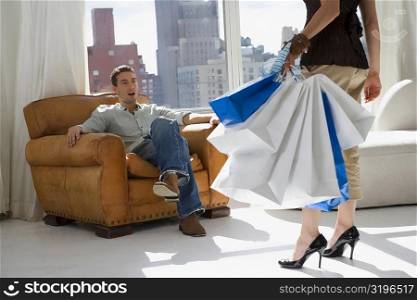 Low section view of a young woman carrying shopping bags with a young man sitting in an armchair