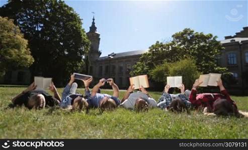 Low view of busy students lying on their backs on park lawn, reading books, browsing touchpad and smartphone while preparing for exams on university campus. Dolly shot. Group of college freinds learning together on green grass.
