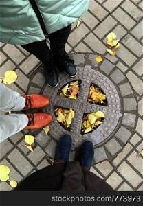 Luke with the inscription Sewerage of Leningrad. Top view of the pavement tile with autumn leaves. Feet in the shoes of two women and one man. St. Petersburg, Russia, tourism.. Luke with the inscription Sewerage of Leningrad.