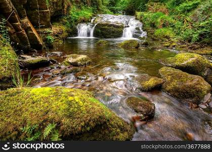 Lush green forest scene with long exposure blurred waterfall flowing through and over rocks covered in lichen and moss