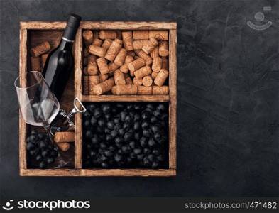 Luxury bottle of red wine and empty glass with dark grapes with corks and corkscrew inside vintage wooden box on black stone background. Space for text
