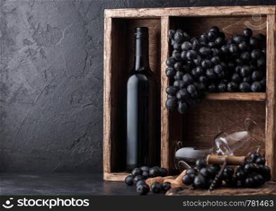 Luxury bottle of red wine and empty glasses with dark grapes inside vintage wooden box on black stone background. Corks and corkscrew on black board.