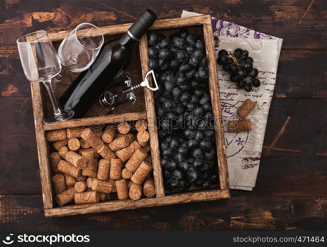 Luxury bottle of red wine and empty glasses with dark grapes with corks and corkscrew inside vintage wooden box on dark wooden background with linen towel.