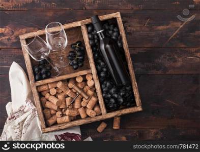 Luxury bottle of red wine and empty glasses with dark grapes with corks and corkscrew inside vintage wooden box on dark wooden background with linen towel.