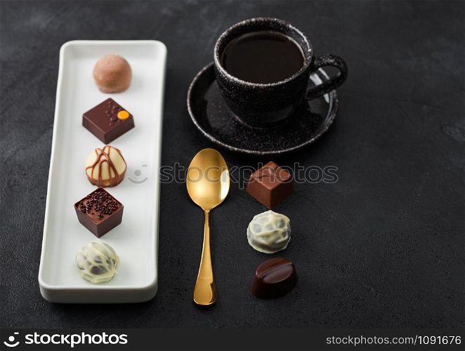 Luxury Chocolate candies in white porcelain plate with cup of black coffee and golden spoon on dark background. Top view