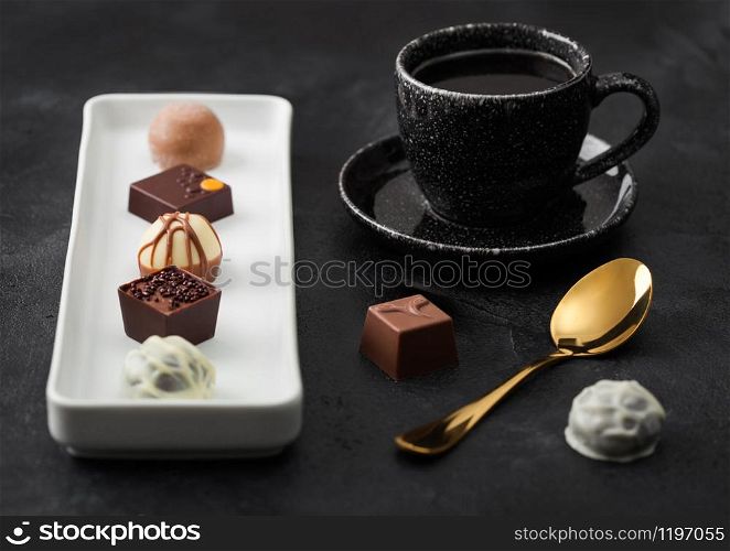 Luxury Chocolate candies in white porcelain plate with cup of black coffee and golden spoon on dark background.