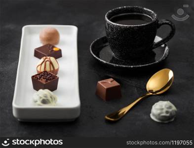 Luxury Chocolate candies in white porcelain plate with cup of black coffee and golden spoon on dark background.