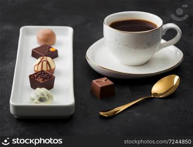 Luxury Chocolate candies in white porcelain plate with cup of black coffee and golden spoon on dark background. Top view
