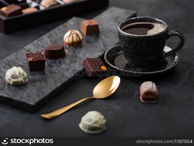 Luxury Chocolate candies selection with cup of black coffee and golden spoon on black marble board and dark background.