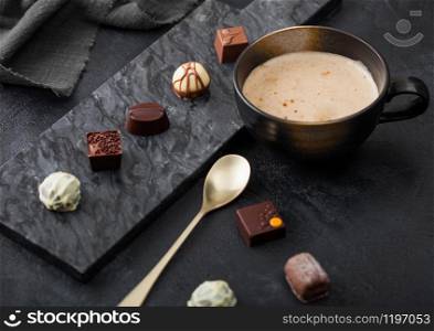 Luxury Chocolate candies selection with cup of cappuccino coffee and dessert spoon on black marble board and dark background.