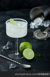 Luxury glass of Margarita cocktail with fresh limes and bar spoon with ice cubes in shaker on black table background.