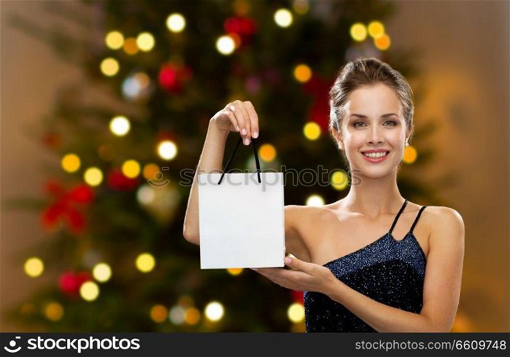 luxury, holidays and sale concept - smiling woman with white blank shopping bag or gift over christmas tree lights background. woman with white shopping bag on christmas