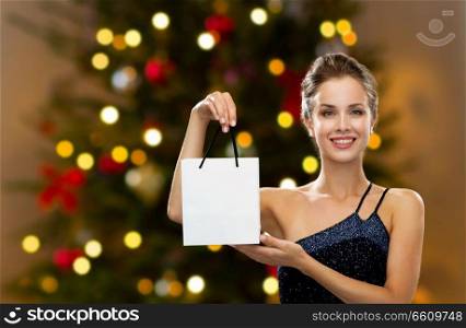 luxury, holidays and sale concept - smiling woman with white blank shopping bag or gift over christmas tree lights background. woman with white shopping bag on christmas