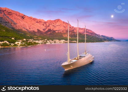 Luxury yacht and blue sea at sunset in summer. Aerial view of big modern sail boat. Top view of beautiful futuristic yacht, water, city, green trees, mountains, purple sky with moon. Adriatic sea