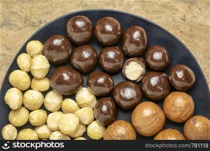 macadamia nuts in shells, shelled and dipped in dark chocolate on a black plate