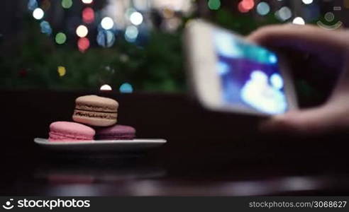 Macaroons on the table with following focus on female hand taking a picture of dessert using phone camera.
