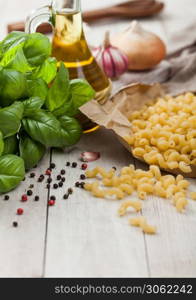 Maccheroni elbows classic raw pasta in brown paper on light wooden table background with basil and oil, pepper and onion.