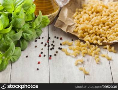 Maccheroni elbows classic raw pasta in brown paper on light wooden table background with basil and oil.