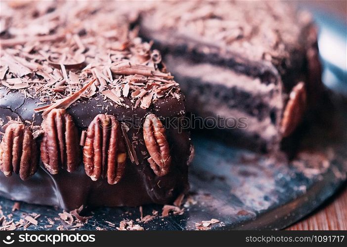Macro view of delicious dark chocolate cake with chips, beautiful icing and pecan nuts on the side on the metal dish. Selective focus. Luxurious glaze. Image for menu or confectionery catalog