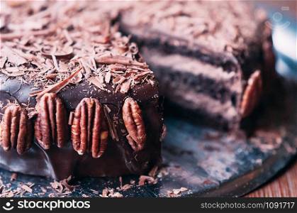 Macro view of delicious dark chocolate cake with chips, beautiful icing and pecan nuts on the side on the metal dish. Selective focus. Luxurious glaze. Image for menu or confectionery catalog