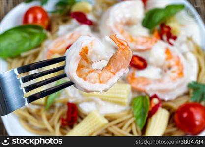 Macro view of stainless steel fork holding single shrimp with Alfredo Pasta dinner placed on white plate in background