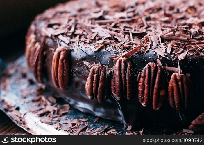 Macro view of whole dark chocolate cake with beautiful icing, chips and pecan nuts on the side on the metal dish. Selective focus. Luxurious glaze. Image for menu or confectionery catalog