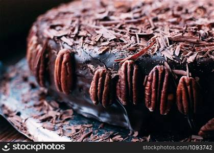 Macro view of whole dark chocolate cake with beautiful icing, chips and pecan nuts on the side on the metal dish. Selective focus. Luxurious glaze. Image for menu or confectionery catalog