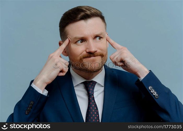 Mad angry businessman in suit holding fingers on his temples while receiving bad news, emotional male entrepreneur feeling annoyed and looking aside while standing against light steel blue background. Angry upset businessman thinking about business problems, trying to find solution