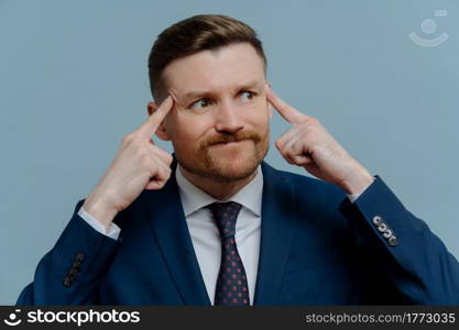 Mad angry businessman in suit holding fingers on his temples while receiving bad news, emotional male entrepreneur feeling annoyed and looking aside while standing against light steel blue background. Angry upset businessman thinking about business problems, trying to find solution