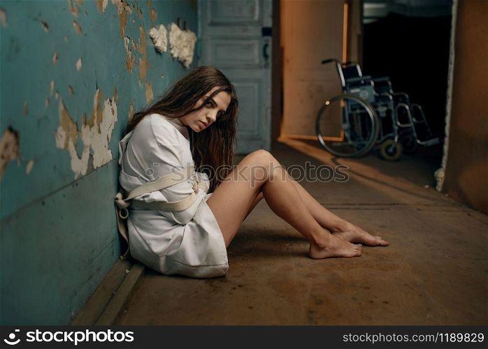 Mad female patient in straitjacket sitting on the floor, mental hospital. Woman in strait jacket undergoing treatment in clinic for the mentally ill, insanity. Mad patient in straitjacket sitting on the floor