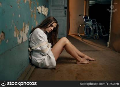 Mad female patient in straitjacket sitting on the floor, mental hospital. Woman in strait jacket undergoing treatment in clinic for the mentally ill, insanity. Mad patient in straitjacket sitting on the floor