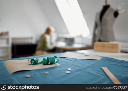 Maesuring tape on the table, jacket on mannequin and seamstress at the sewing machine, workshop interior on background. Dressmaking occupation, handmade tailoring business, handicraft hobby. Maesuring tape on the table, workshop interior
