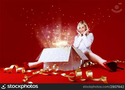 Magic surprise for a pretty blonde! Charming girl in white dress spread shot. Gift box in center. Light beams and stars coming from the box. Red background. Amazing face expression. Sense of holiday.