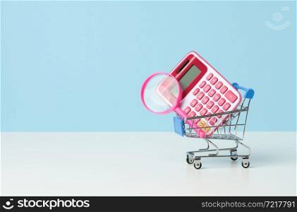 magnifier and pink plastic calculator in a miniature pink trolley on a blue background. Budget planning concept, savings counting, cost control
