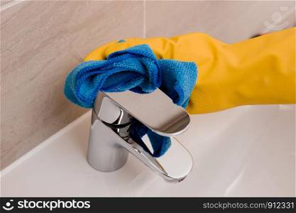 Maid with rubber glove cleaning tap and sink. Housekeeping scrubbing and polishing silver tap in bathroom.