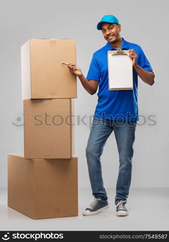 mail service and shipment concept - happy indian delivery man with boxes and clipboard in blue uniform over grey background. indian delivery man with boxes and clipboard