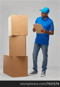 mail service and shipment concept - happy indian delivery man with boxes and clipboard in blue uniform over grey background. indian delivery man with boxes and clipboard