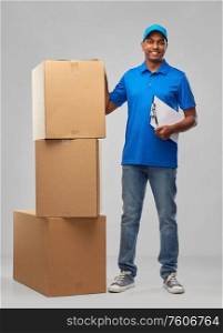 mail service and shipment concept - happy indian delivery man with boxes and clipboard in blue uniform over grey background. indian delivery man with boxes and clipboard