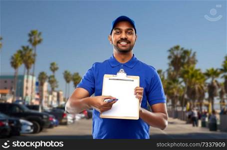 mail service and shipment concept - happy indian delivery man with clipboard in blue uniform over venice beach background in california. happy indian delivery man with clipboard in blue