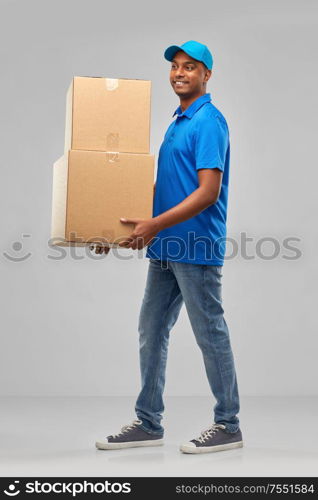 mail service and shipment concept - happy indian delivery man with parcel boxes in blue uniform over grey background. happy indian delivery man with parcel boxes