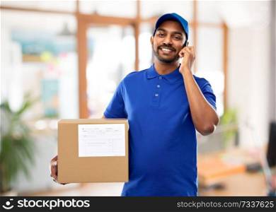 mail service, communication and shipment concept - happy indian delivery man with smartphone and parcel box in blue uniform over office background. indian delivery man with smartphone and parcel box