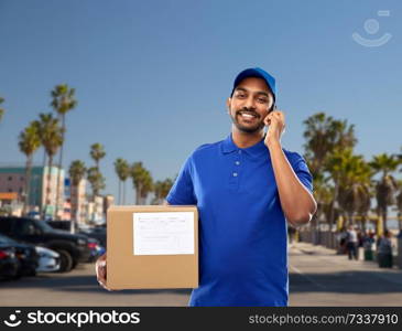 mail service, communication and shipment concept - happy indian delivery man with smartphone and parcel box in blue uniform over venice beach background in california. indian delivery man with smartphone and parcel box