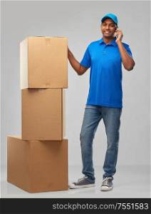 mail service, communication and shipment concept - happy indian delivery man with smartphone and parcel boxes in blue uniform over grey background. indian delivery man with boxes calls on smartphone