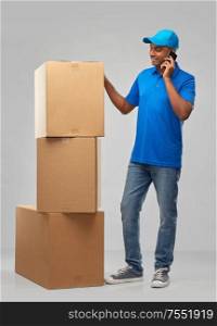 mail service, communication and shipment concept - happy indian delivery man with smartphone and parcel boxes in blue uniform over grey background. indian delivery man with boxes calls on smartphone