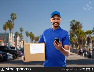mail service, technology and shipment concept - happy indian delivery man with smartphone and parcel box in blue uniform over venice beach background in california. indian delivery man with smartphone and parcel box